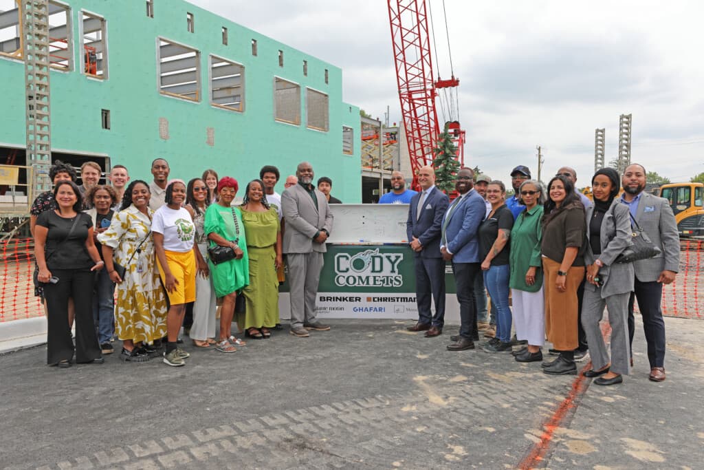 About 30 people pose with a signed steel beam and a Cody Comets banner in front of the new Cody High School building under construction, with a red crane behind them.