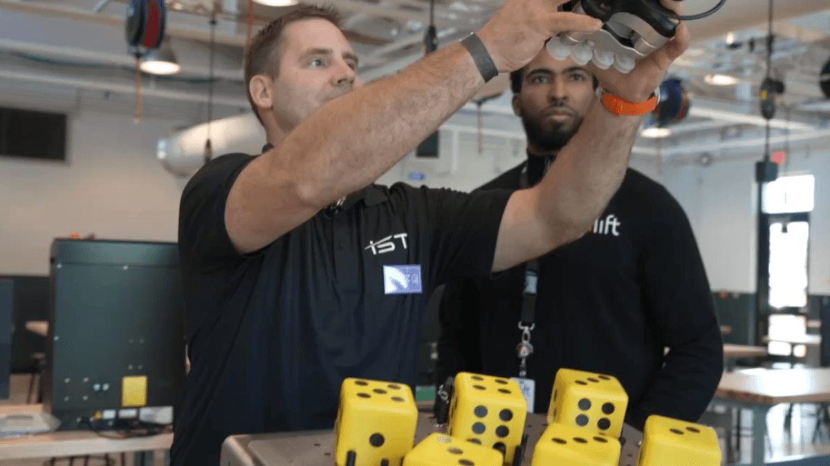 A man in a black polo shirt operates a robotic hand above a tray of yellow foam dice in the new career technical education annex.